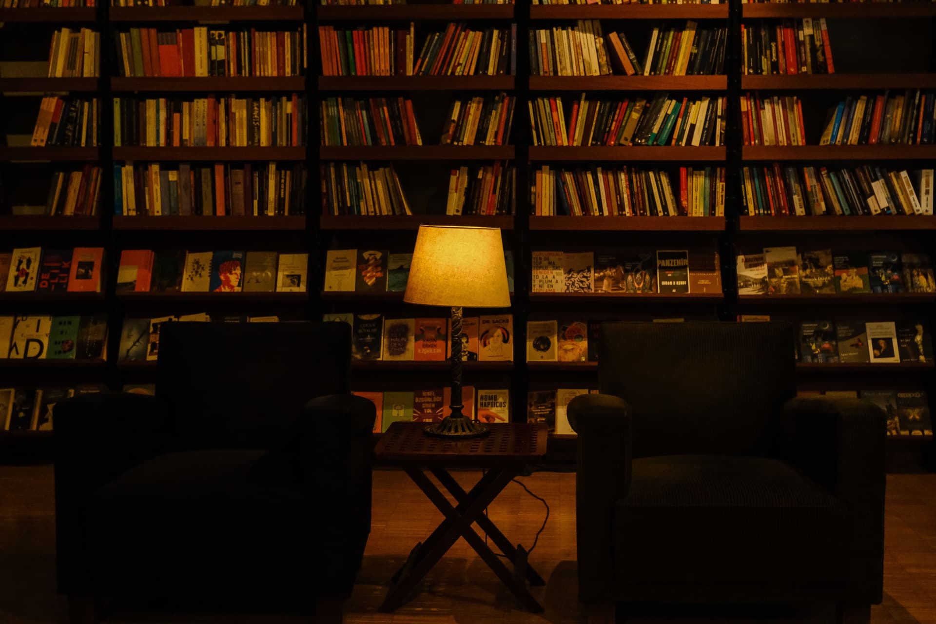 A dimly lit reading room with full bookshelves, two dark armchairs and a warm lamp between them