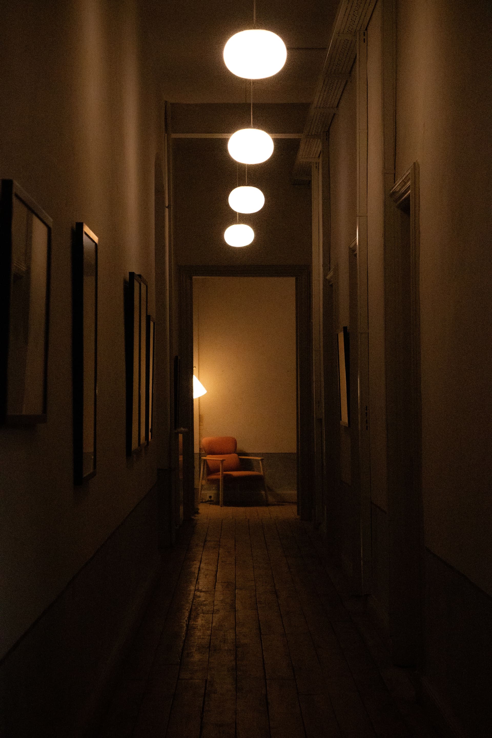 A dim hallway with wooden floors leading toward a glowing room with a chair at the far end