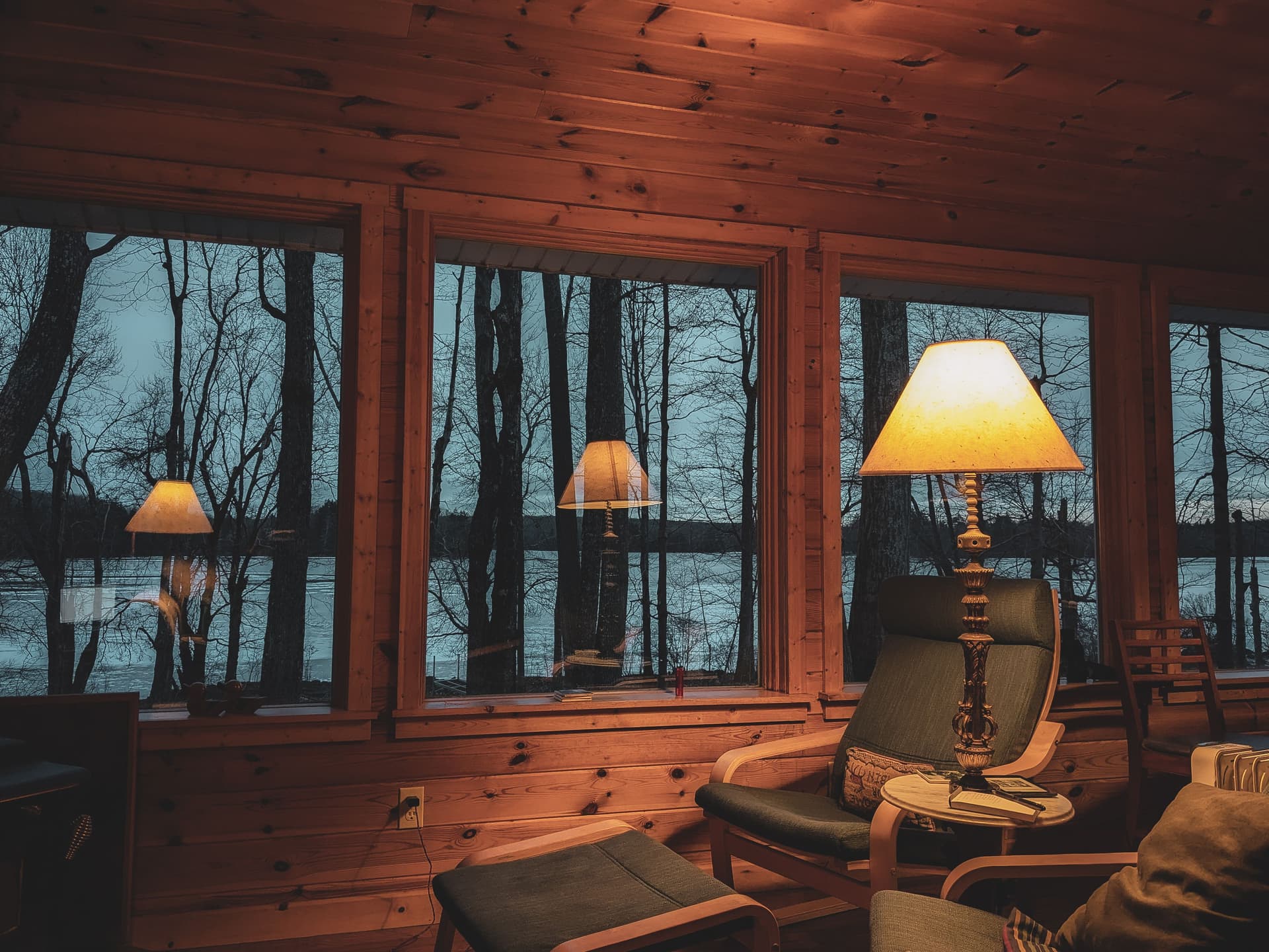 A dim cabin interior with wood-paneled walls, lit lamps and a chair by windows that look out on a lake at dusk